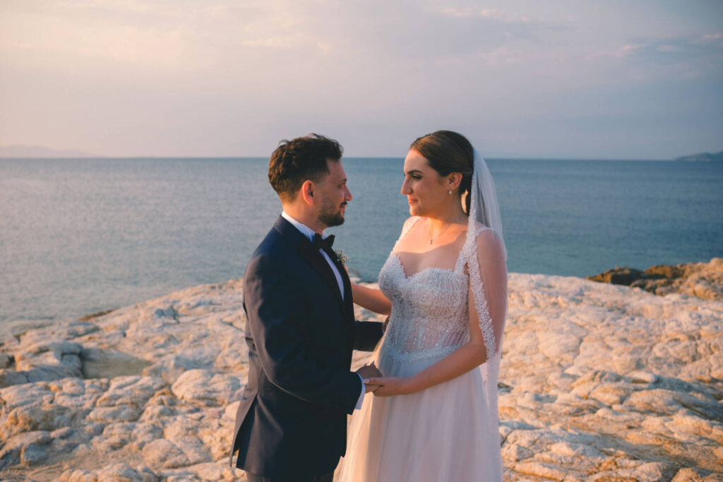 Το ζευγάρι κρατιέται από τα χέρια στα βράχια με θέα στη θάλασσα, στο ηλιοβασίλεμα